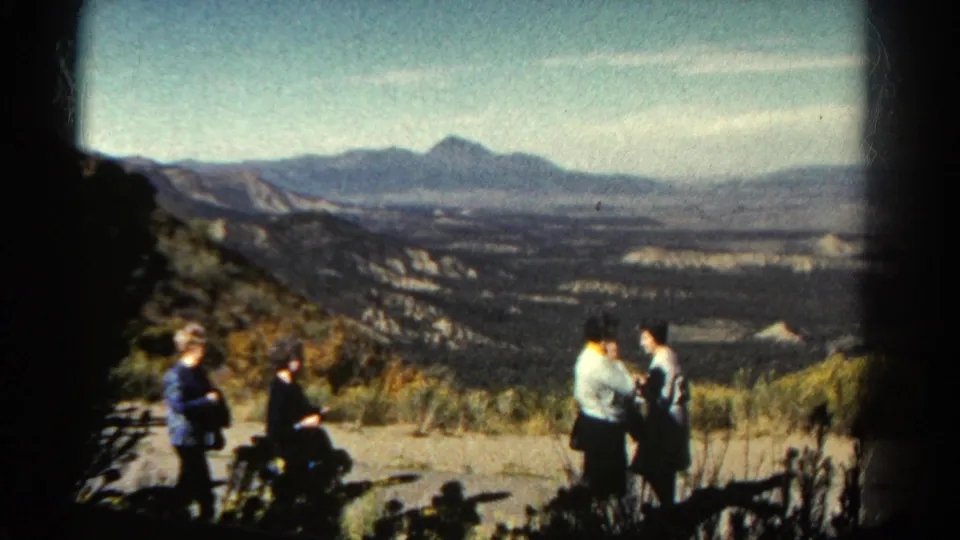 Two adult women stand together near the right edge of a Colorado overlook while a seated woman and another figure are visible at left against the valley.