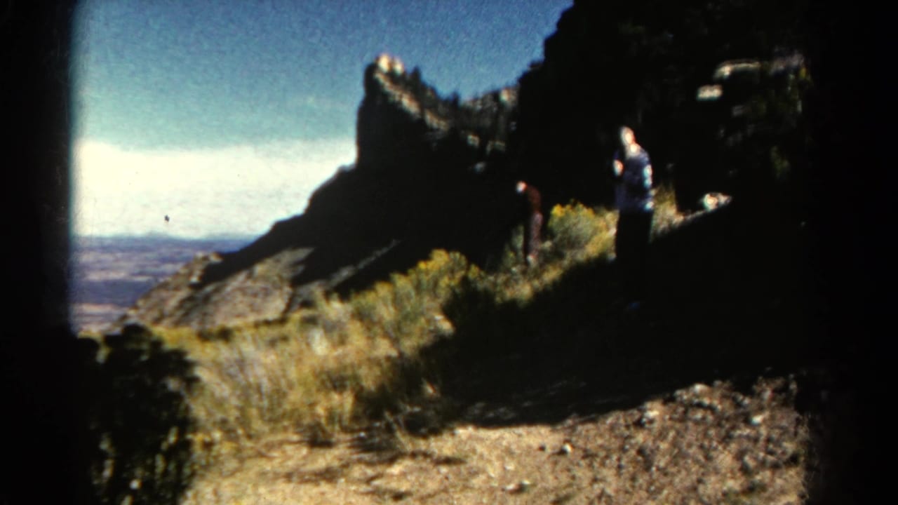 Two visitors stand on a rocky overlook trail beside dry grass and dark cliff faces.
