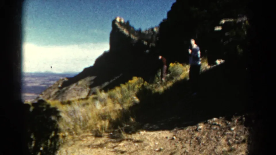 1967 Colorado 8mm frame showing two visitors on a rocky overlook trail with dry grass, dark rock formations, and distant valley haze.