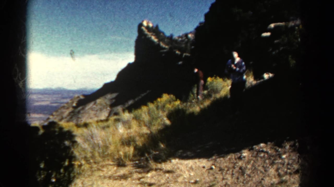 1967 Colorado overlook view with visitors near a dirt trail, dry brush, steep rock formations, and a broad valley in the distance.