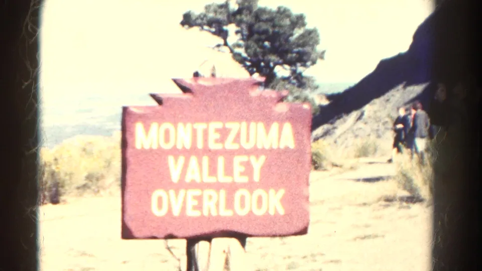 1967 Colorado 8mm frame with a red Montezuma Valley Overlook sign, trail, tree, rock slope, and visitors near the viewpoint.