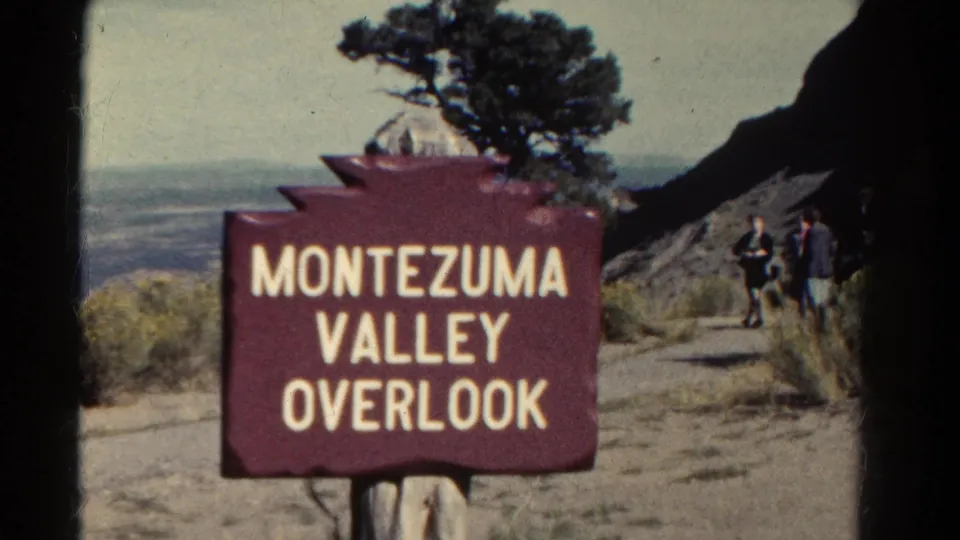 1967 Colorado frame centered on the Montezuma Valley Overlook sign with visitors, a tree, rocky slope, and hazy valley beyond.