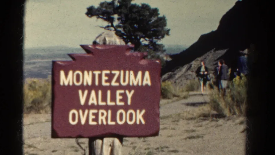 1967 8mm Colorado frame of the Montezuma Valley Overlook sign with distant visitors, tree, rock slope, and valley view.