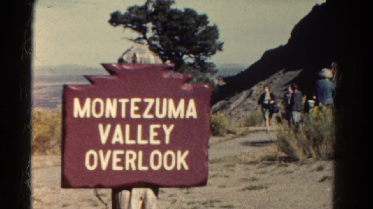 Montezuma Valley Overlook sign beside a dirt path with tourists and a cliff in the background.