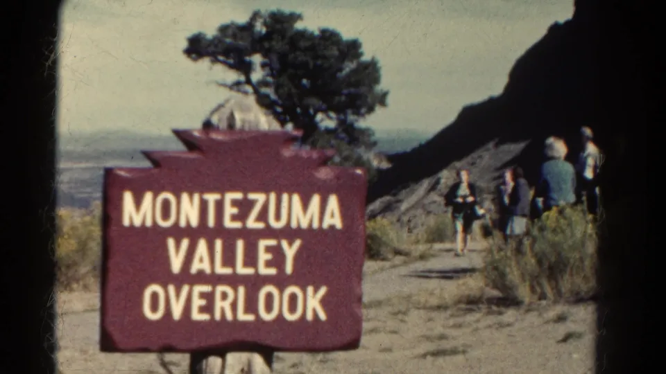 1967 Colorado 8mm frame showing the Montezuma Valley Overlook sign with tourists on the path and desert vegetation around the viewpoint.