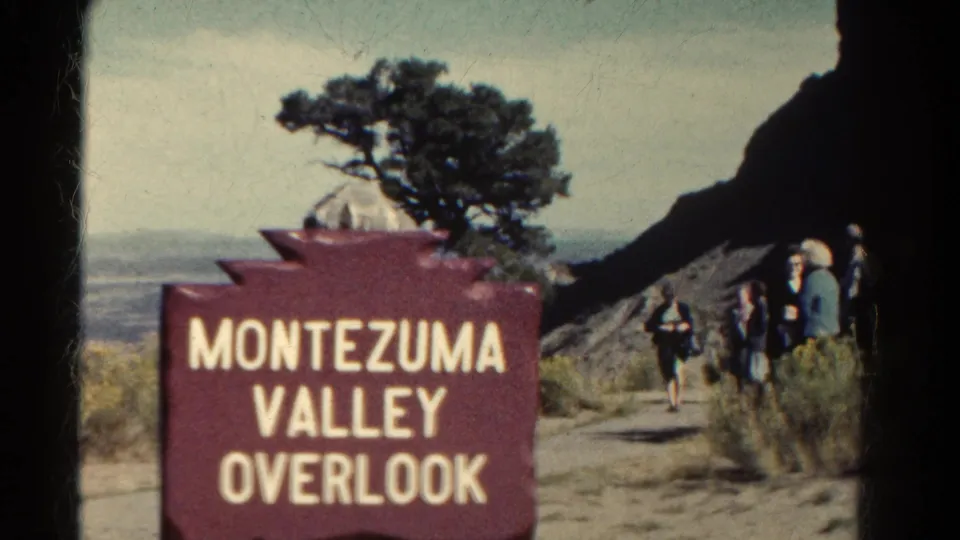 1967 Colorado home-movie still of the overlook sign and tourists at the path edge, with rugged rock, brush, and open valley beyond.