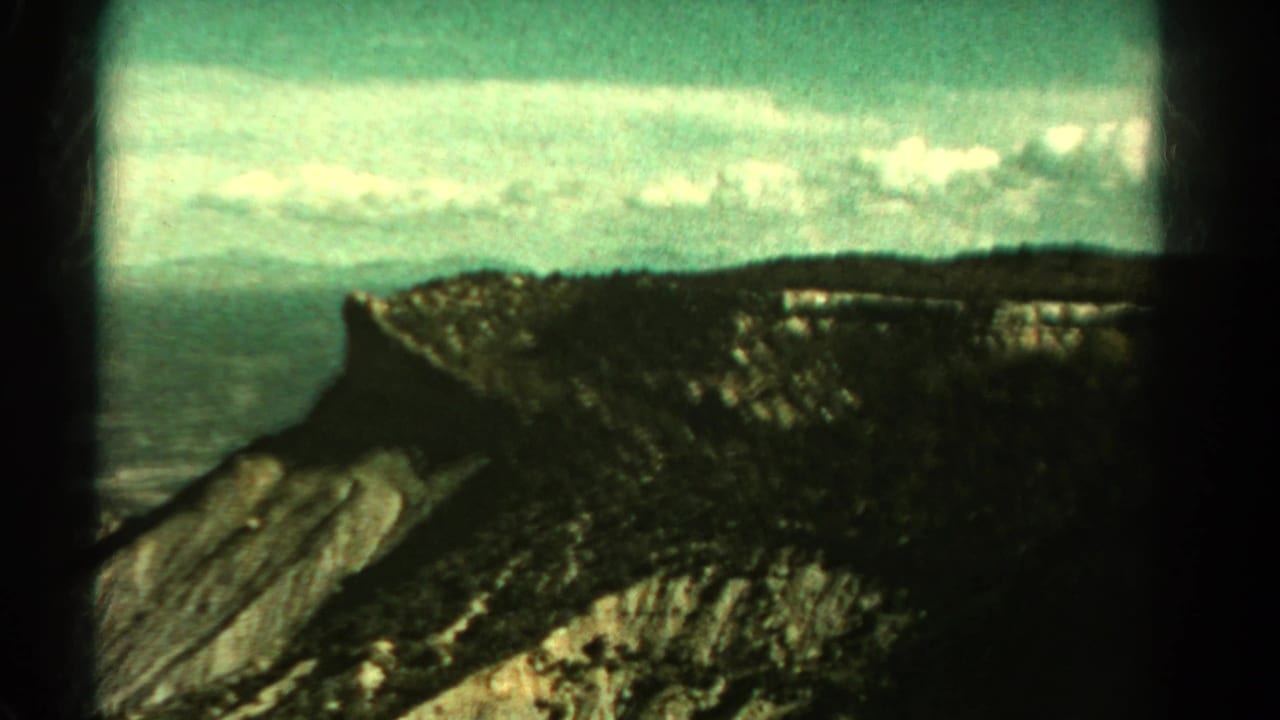 Aerial-looking 1967 Colorado mountain view with a steep cliff, forested ridge, pale sky, and visible 8mm frame edges.