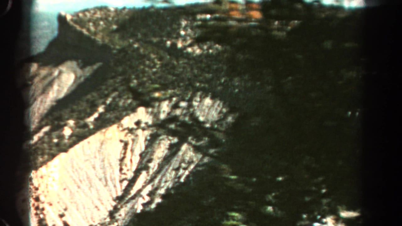 1967 Colorado 8mm frame showing dark forested slopes, layered cliffs, and a bright exposed quarry face across a mountain valley.