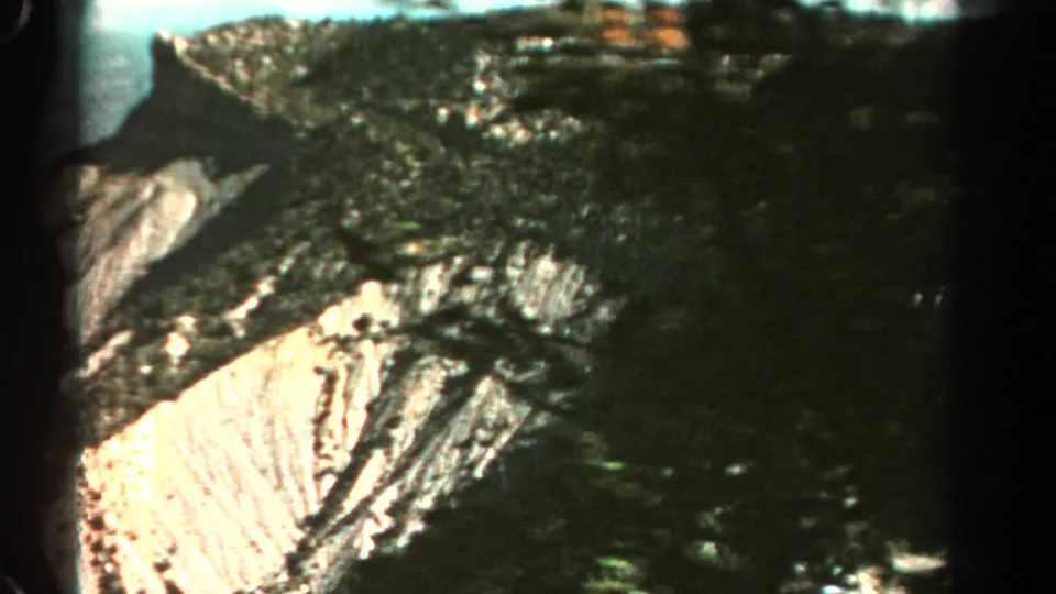 1967 Colorado 8mm frame showing dark forested slopes, layered cliffs, and a bright exposed quarry face across a mountain valley.