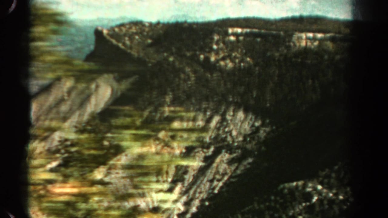 1967 Colorado home-movie view across a rugged valley with forested cliffs, exposed rock faces, and a distant ridgeline under bright sky.
