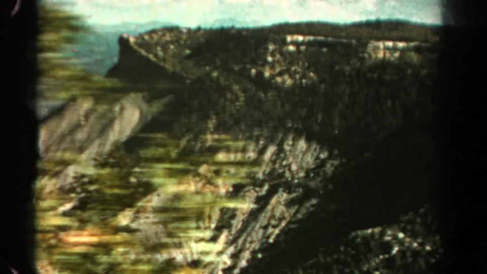 1967 Colorado home-movie view across a rugged valley with forested cliffs, exposed rock faces, and a distant ridgeline under bright sky.
