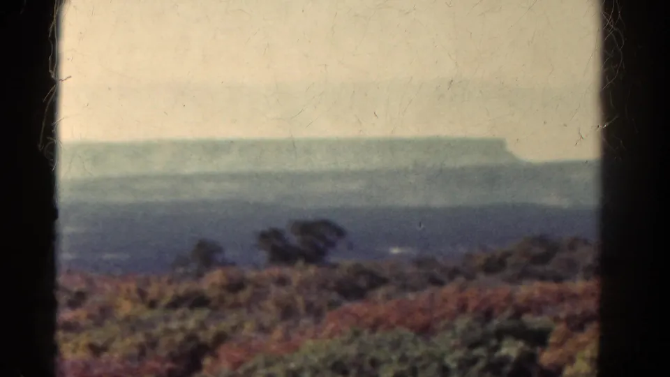 1967 Colorado landscape frame with muted brush, layered haze, and a distant mesa-like plateau beneath a washed pale sky.