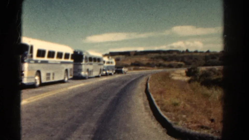 A 1967 Colorado home-movie frame shows the bus convoy receding around a road curve under bright daylight.