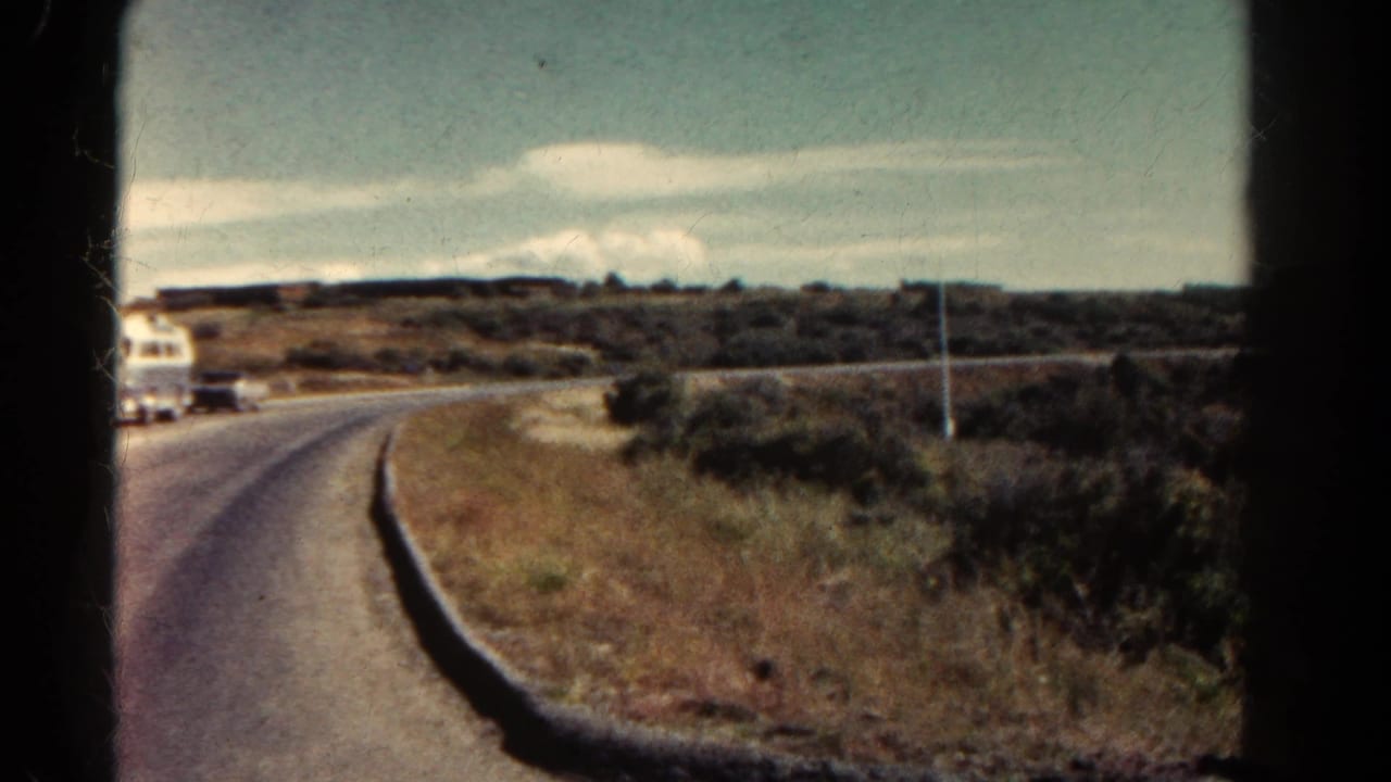 1967 Colorado roadside view with a white bus at the edge of frame, a paved road, scrubby vegetation, low hills, and pale sky.