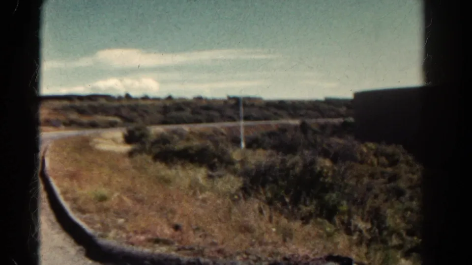 1967 Colorado 8mm frame showing roadside shrubs, dry grass, a curved road edge, low hills, and a dark structure at the right.