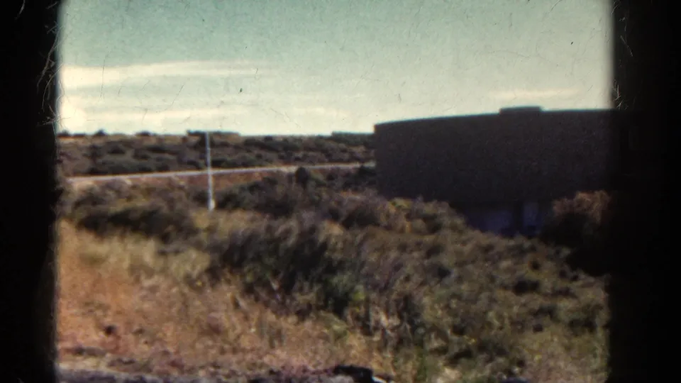 1967 Colorado roadside frame with a dark building-like structure, scrub vegetation, paved road, low hills, and pale sky.