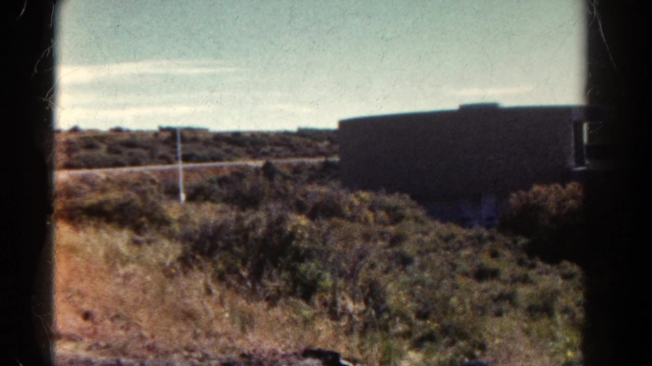 1967 Colorado 8mm frame showing roadside terrain, a low structure, pale sky, and dark film borders before the lodge sign close-up.
