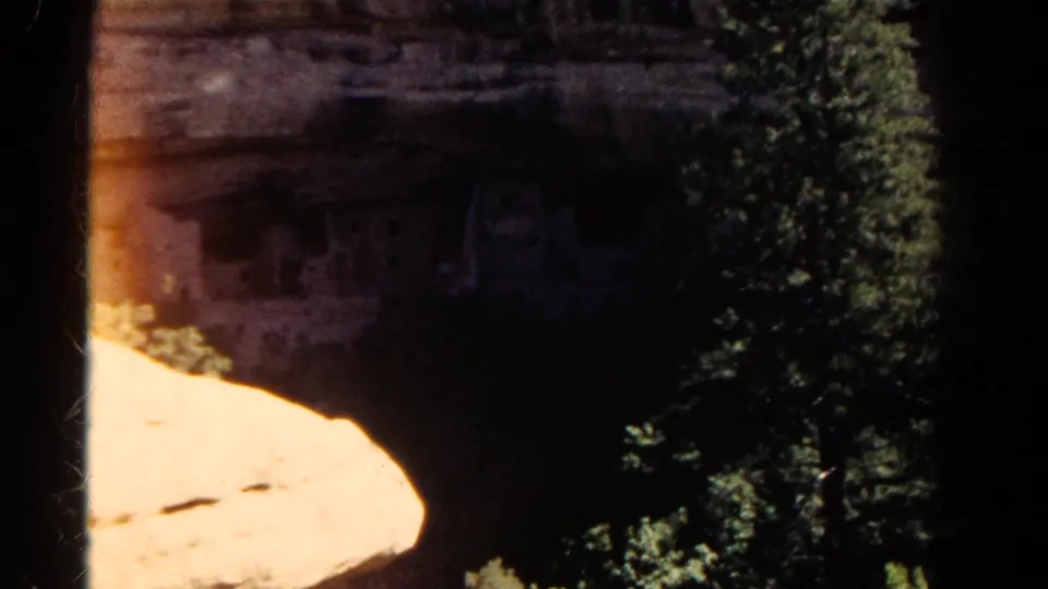 1967 Colorado 8mm frame showing pine trees partly blocking stone ruins set into a shaded cliff alcove.