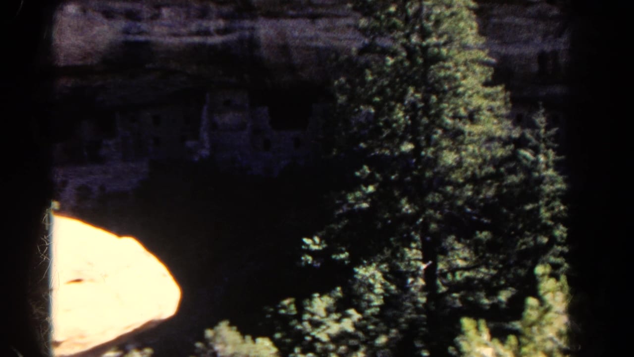 Colorado home-movie frame with cliffside ruins in shadow behind pine trees and a bright sunlit rock shelf.