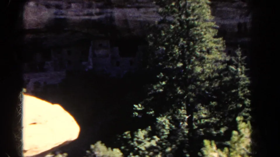 Colorado home-movie frame with cliffside ruins in shadow behind pine trees and a bright sunlit rock shelf.