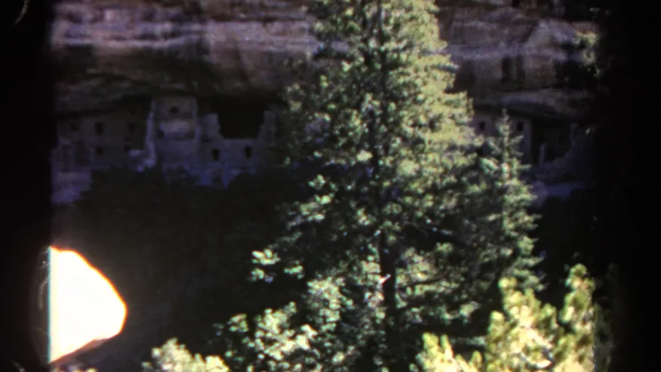 1967 Colorado 8mm still with bright evergreens in front of shadowed stone ruins beneath a layered cliff.
