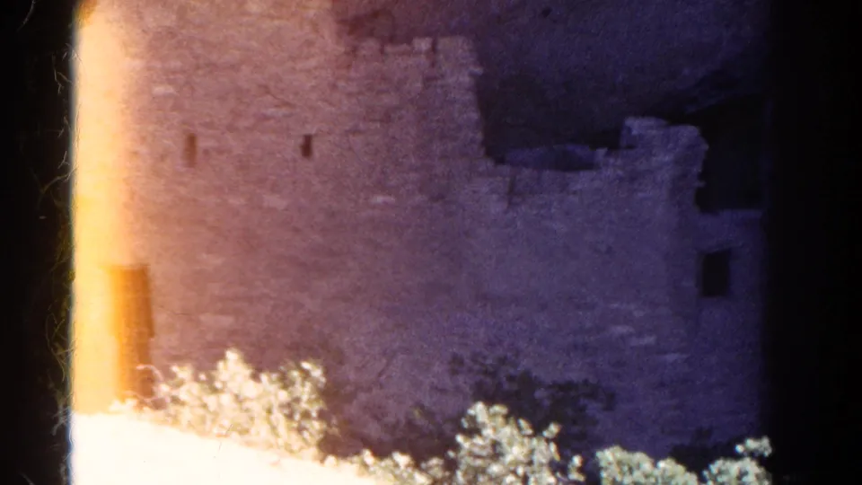 Grainy 8mm frame of Cliff Palace masonry, a dark doorway, shrubs, and a vertical light flare at the left edge.