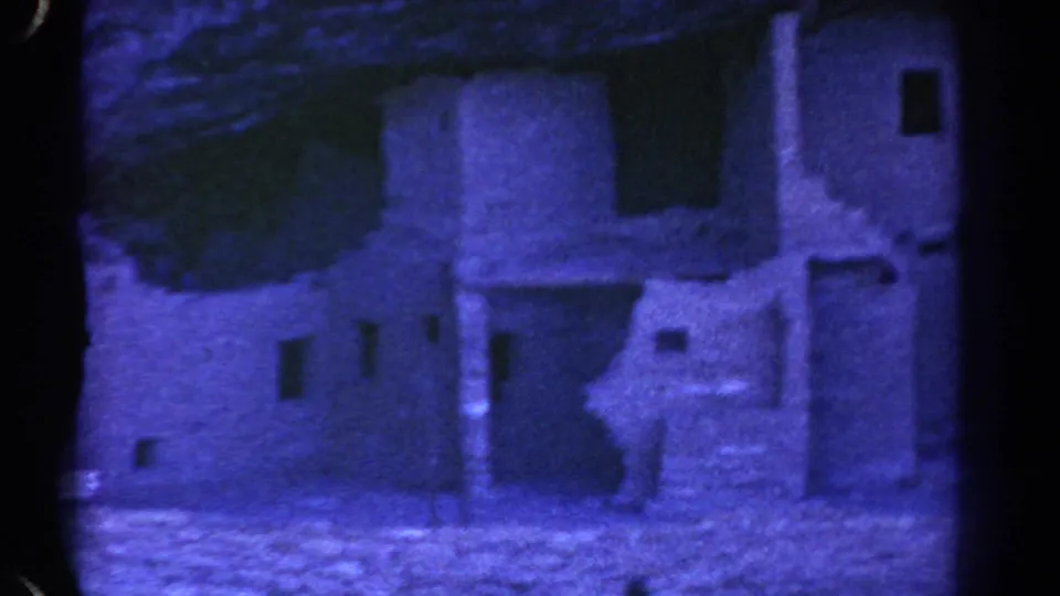 1967 Colorado travel-film frame of a stone ruin facade with a doorway, small openings, signs and a strong blue tint.
