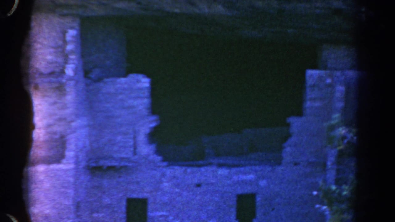 1967 Colorado 8mm frame showing a weathered cliff-side ruin facade with small openings, seen through dark tree edges and blue-shifted light.