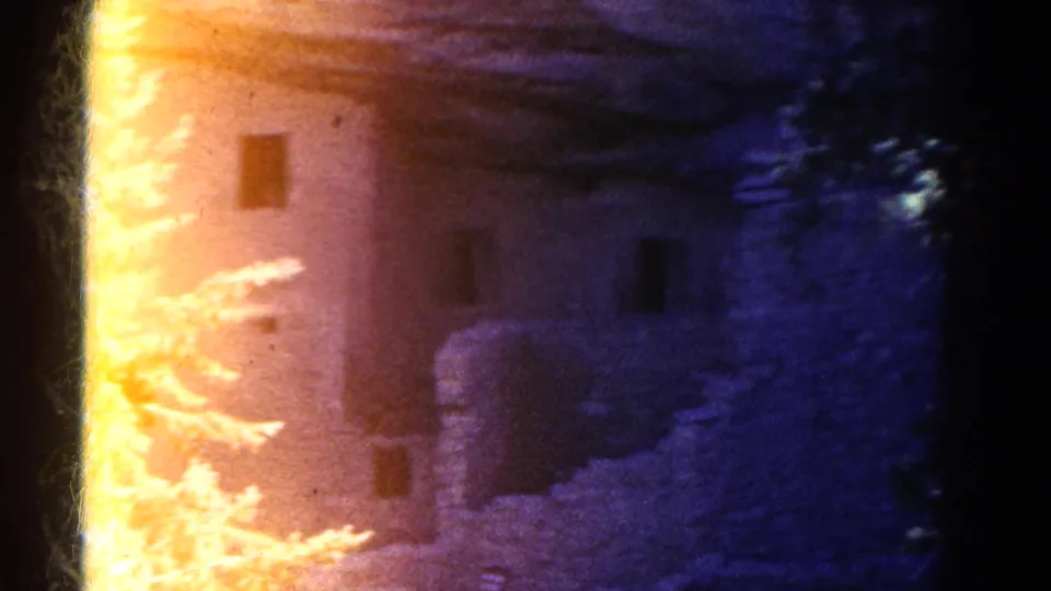 1967 Colorado 8mm frame showing a warm-lit cliff ruin wall with small dark openings, shadowed rock, and trees at the edge of the shot.