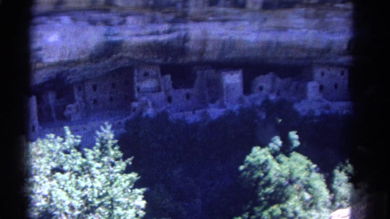 1967 Colorado 8mm frame showing ancient cliff dwellings set into a rock alcove, partly obscured by bright trees in the foreground.