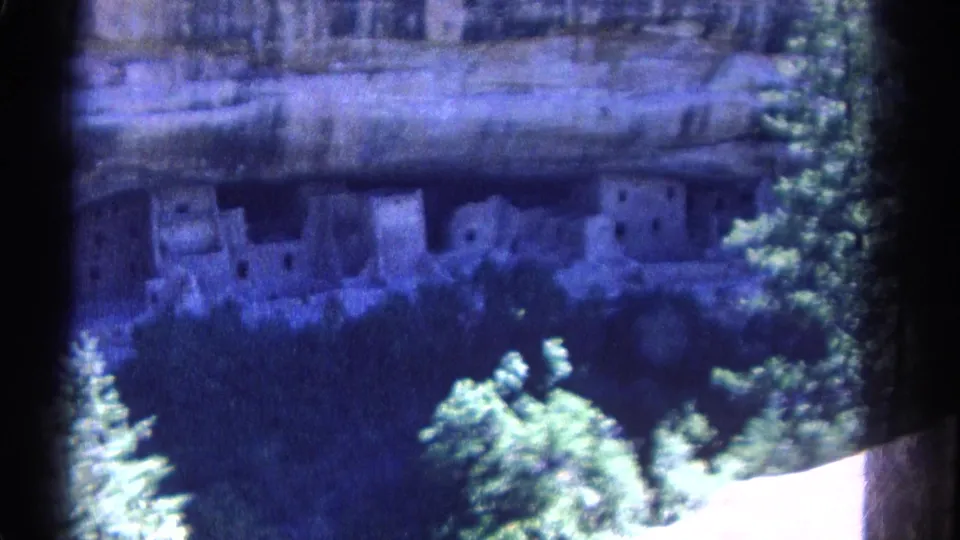 1967 Colorado travel frame showing cliff dwellings across a shaded canyon, with evergreen trees below and a soft blue-purple film cast.