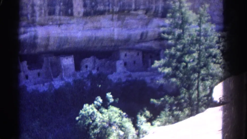 Final 1967 Colorado 8mm frame of cliff dwellings in a rock alcove, framed by trees, canyon shadow, and bright pale rock at lower right.