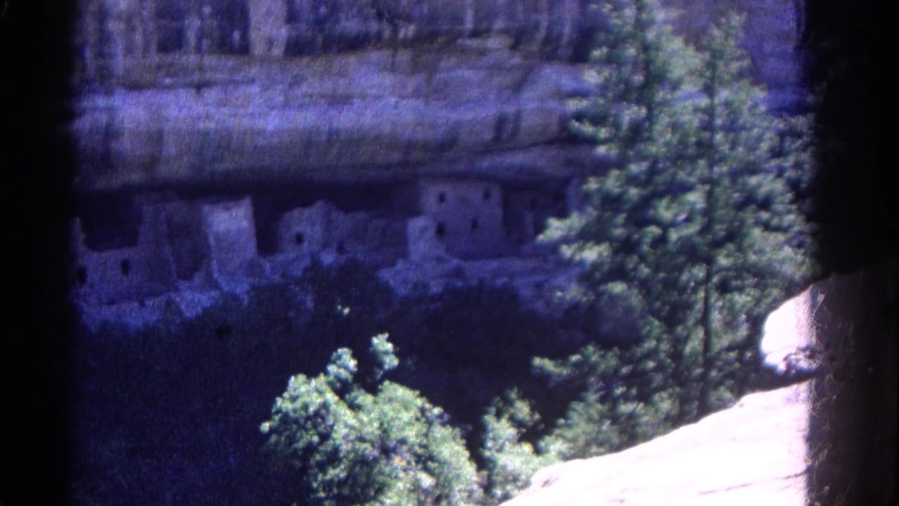 1967 Colorado 8mm frame showing cliff dwellings in a shaded rock alcove, with green trees and a sunlit stone ledge nearby.