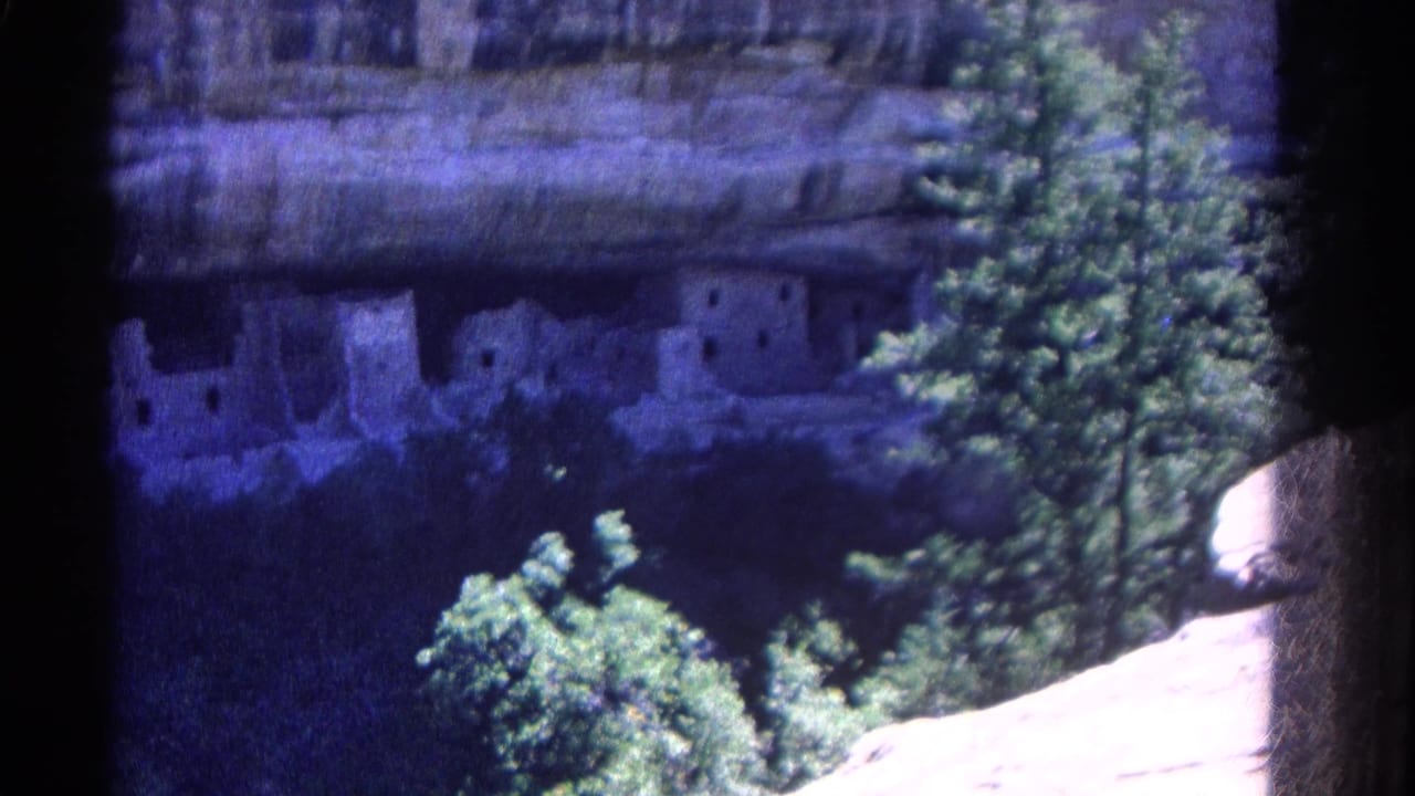 1967 Colorado home-movie view of cliff dwellings across a canyon-like overlook, framed by trees and bright rock.