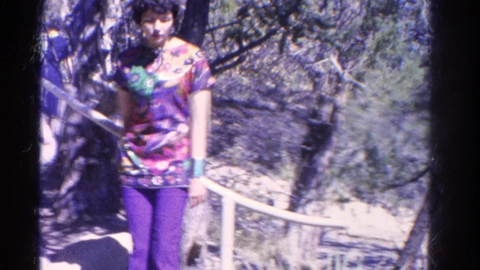 1967 Colorado home-movie frame showing an adult woman by a white overlook railing, wearing a colorful patterned top and purple pants.