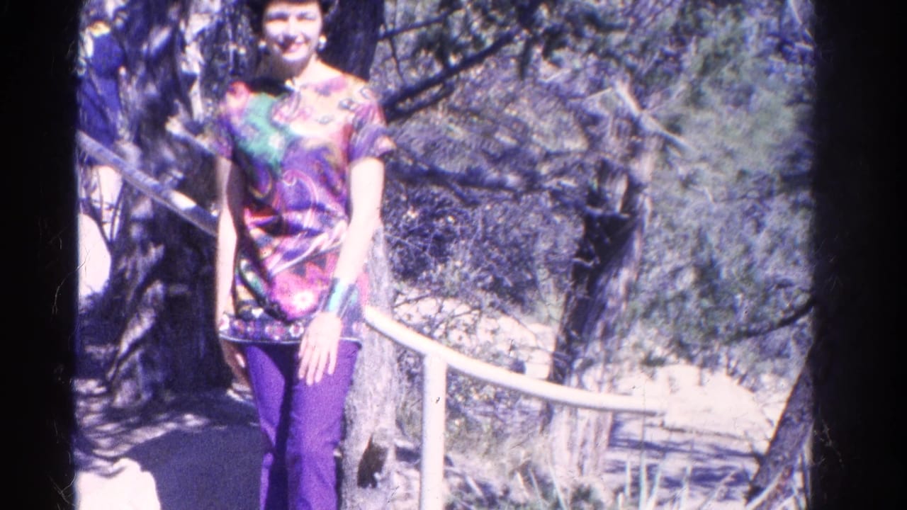 A woman in purple pants smiles toward the camera on a wooded Colorado trail, with a white railing and bright sun patches behind her.