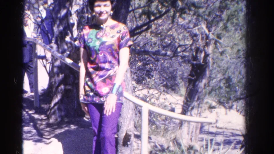 The home-movie frame shows the woman closer to camera in her patterned top and purple pants beside a white railing and trees.