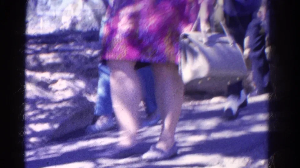 A soft, handheld 8mm frame shows walkers from the waist down on a shaded trail, including a light handbag and colorful tunic.
