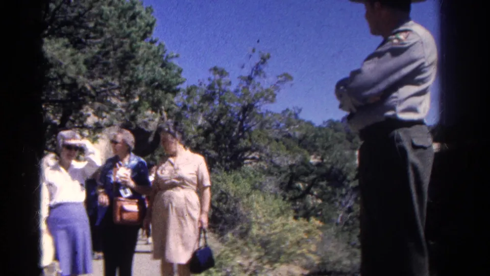 Women in period clothing walk along a Colorado path while a uniformed officer or ranger stands partly silhouetted at the right edge.