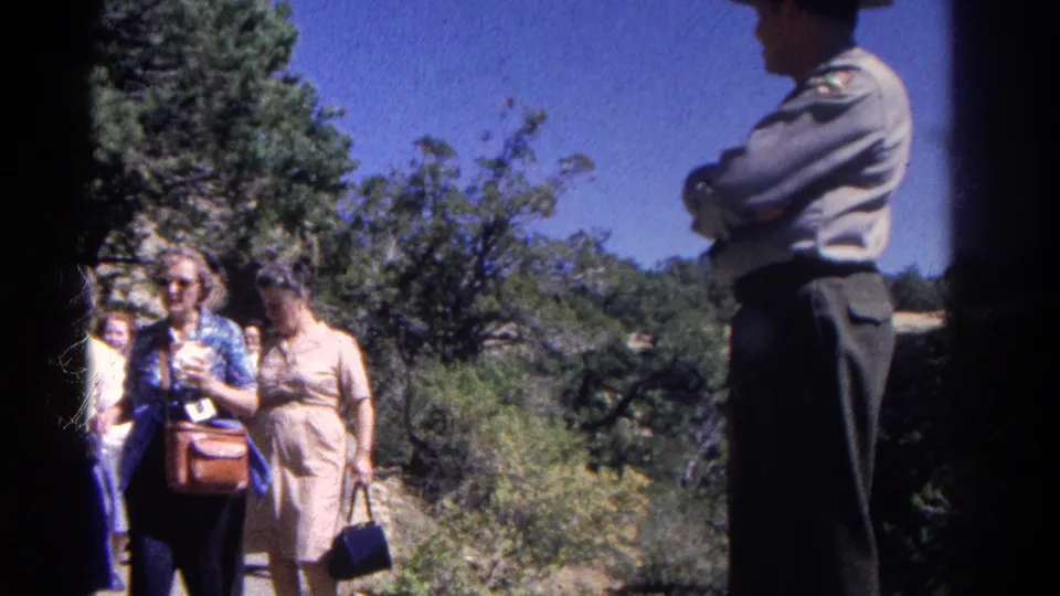 A woman with sunglasses, a blue top, and a shoulder bag passes a uniformed officer or ranger on a sunlit Colorado trail.