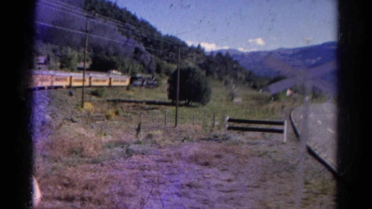 Yellow train cars pass through a rural valley with a road, utility pole, trees, and mountains.