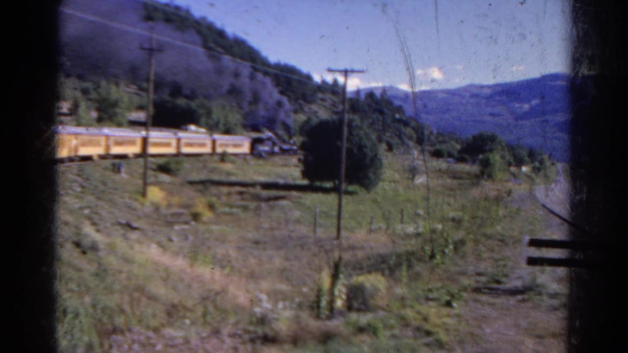Yellow train cars run along the hillside with a utility pole, open field, trees, and mountains.