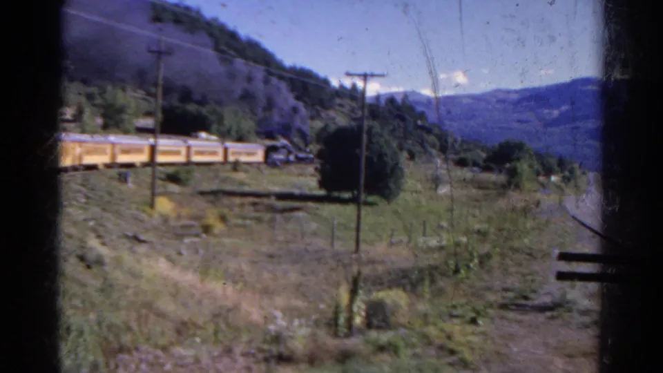 1967 Colorado home-movie frame of yellow rail cars crossing a green rural hillside below distant mountains.