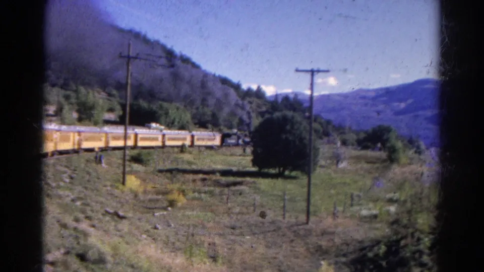 1967 Colorado 8mm still with a yellow train, foreground utility pole, rural slope, trees, and blue mountain backdrop.