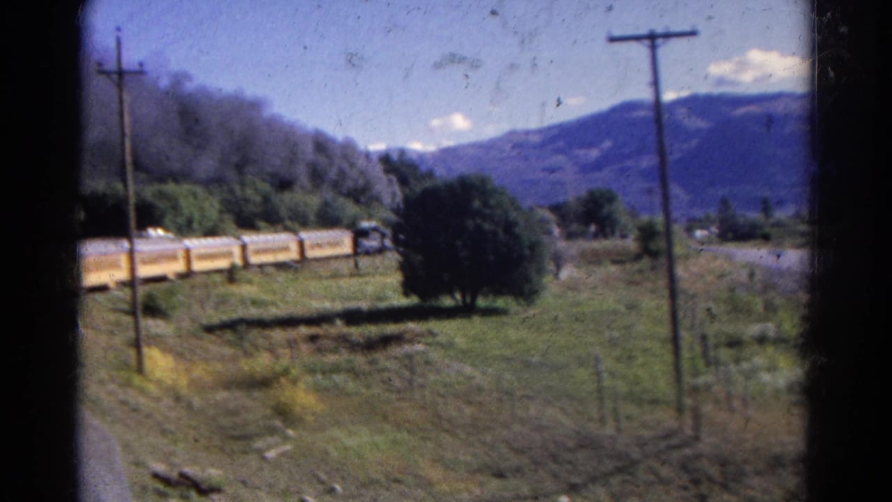 1967 Colorado home-movie frame showing a yellow passenger train crossing open fields with utility poles, trees, and distant mountains.