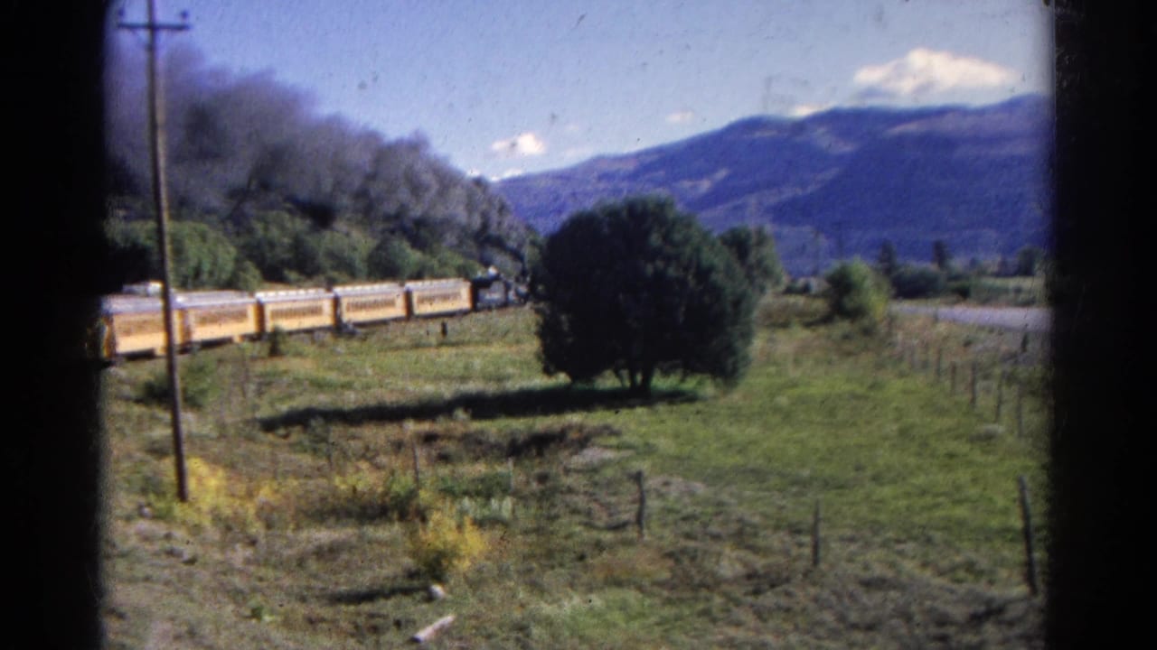 The yellow train continues along the valley track in Colorado, with a smoke plume, trees, fence lines, and mountains behind it.