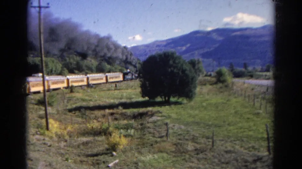 The yellow train continues along the valley track in Colorado, with a smoke plume, trees, fence lines, and mountains behind it.