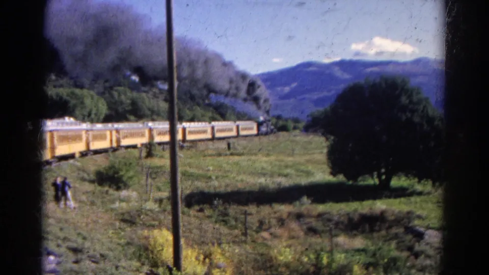 A closer 1967 Colorado view shows the yellow passenger train, dark locomotive smoke, a utility pole, open field, and small distant people.