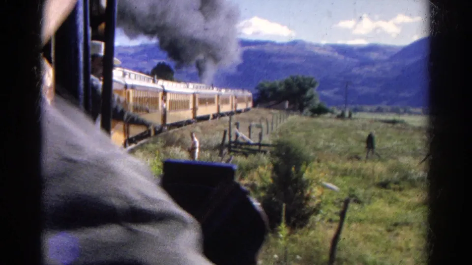 From a passenger viewpoint, the yellow train curves through the Colorado valley while a nearby shoulder and window edge frame the scene.