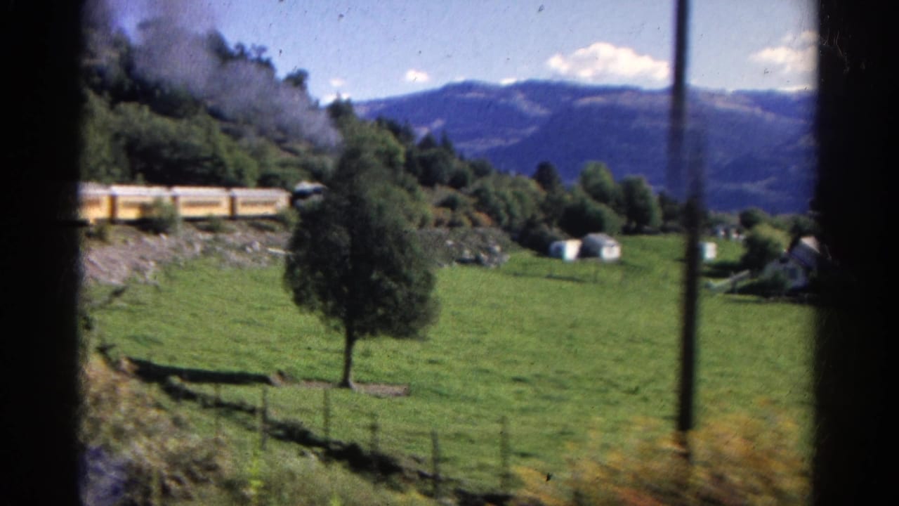 Yellow passenger train and smoke seen across a grassy field with mountains behind.